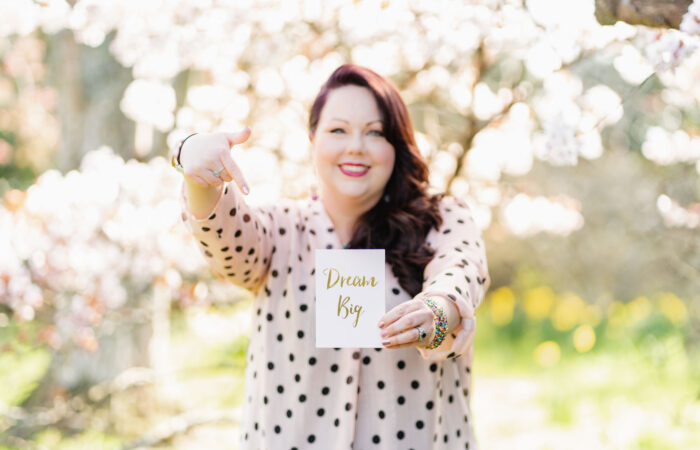 Women with long brown hair wearing pink top with black poka dots smiling at the camera holding a card up that says dream big