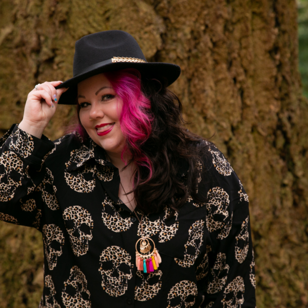 Women smiling in the woods, with pink hair, wearing a black cowboy style hat with gold detail, black shirt with leopard print skulls
