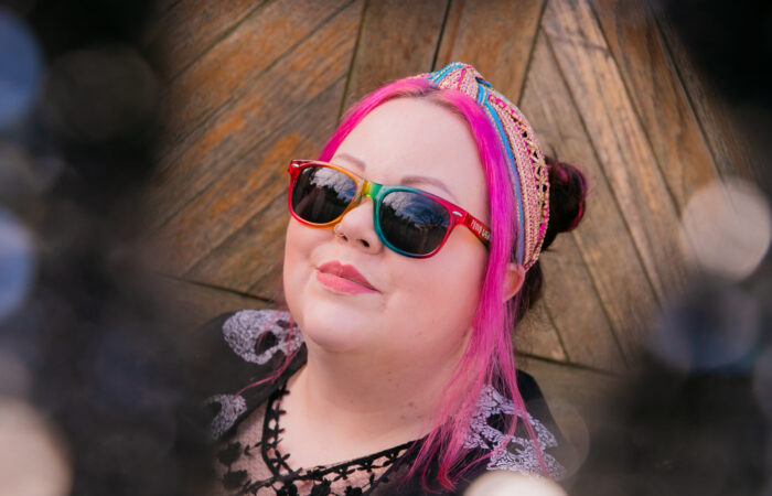 woman with pink hair and rainbow sun glasses smiling into the distance, surrounded by sparkly sequins