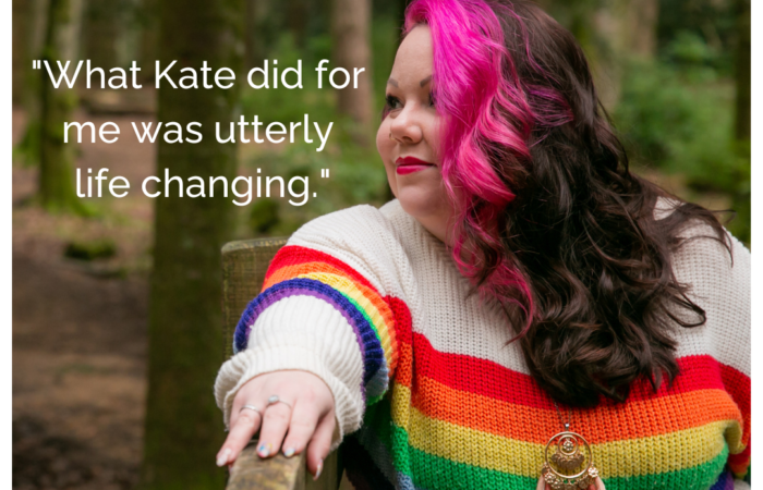Image of a woman with long bright pink hair in a white jumper with rainbow stripes stood in the woods, smiling.