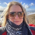 Close up image of a woman smiling with blonde hair, sunglasses and patterned scarf